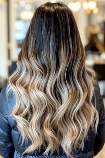 Long Ombre Waves With Dark Roots Hairstyle on a woman with brown hair at roots and blonde hair at the ends, back view in salon.