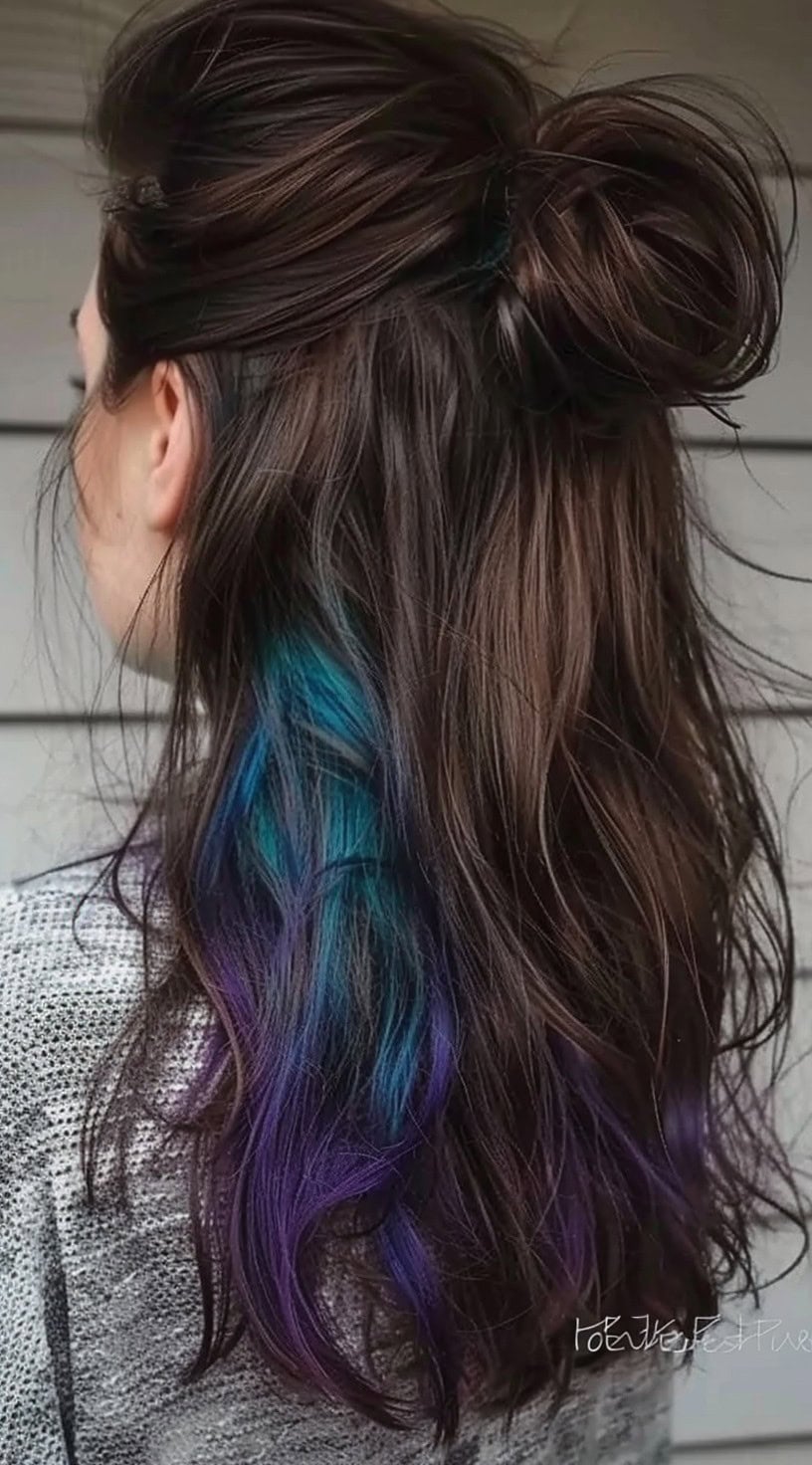 A woman with long, brunette hair styled in a half-up bun, revealing blue and purple highlights underneath.