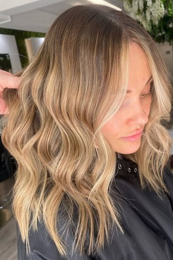 Warm Medium Blonde Balayage Waves on a woman with wavy blonde hair, side view.